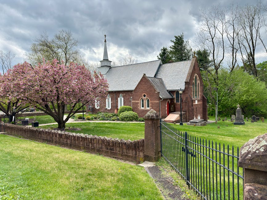 A brick church with a gray slate roof and tall, arched windows is situated within a green graveyard surrounded by mature trees, some with budding leaves and others still bare. In the foreground, a blossoming pink magnolia tree is beside a low brick wall and a black metal gate, which leads into the churchyard. The sky overhead is partly cloudy with patches of dark clouds, indicating an overcast day. The scene captures the peaceful, well-maintained outdoor environment typical of a church setting, with the church building centrally positioned in the image. This setting is relevant for home relocation and moving services, as it illustrates the typical outdoor environment where a removal vehicle might be parked near the property for loading furniture and boxes.