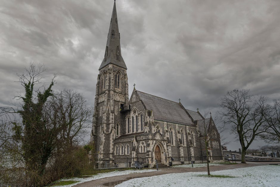 A large historic church with a tall, pointed spire and Gothic architectural features, constructed from stone with light and dark striped patterns, situated outdoors under a cloudy sky. Surrounding the church are leafless trees, some with thick branches and sparse foliage, and a pathway with patches of snow on the ground. In the foreground, there is a fenced area near the entrance with no visible furniture or moving equipment. This scene depicts an exterior view of a church that could be involved in a home relocation process, with no visible boxes, packing materials, or vehicles, but the setting relates to moving services provided by Man with Van St Lukes, particularly in the context of furniture transport or loading procedures at the property.