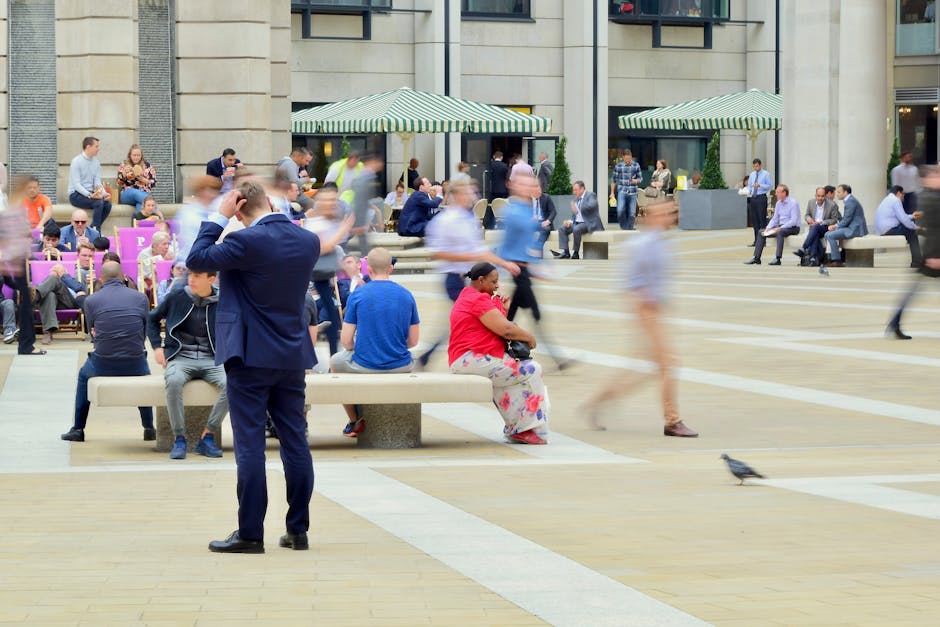 A wide view of a busy public square outside a modern building features numerous people seated on benches and walking across the paved area, with some engaged in conversations and others using their phones. Several individuals are dressed in casual attire, while a man in a dark suit stands with his back to the camera, adjusting his hair or head. In the background, there are striped green and white awnings shading outdoor seating, and additional pedestrians can be seen walking or sitting on steps leading to the building entrance. The scene is set during daytime with natural light illuminating the area, capturing a moment of urban activity linked to home relocation or furniture transport processes, which may involve residents waiting during a house removal service arranged by [COMPANY_NAME], such as Man with Van St Lukes, supporting smooth moving logistics and packing and moving operations.
