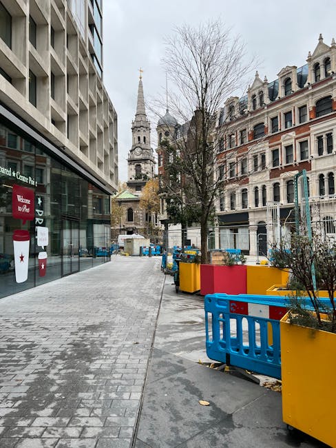 A wide urban street scene during daytime showing a narrow paved walkway adjacent to modern and historic buildings. On the left, a contemporary office or retail building features glass windows and a sign. On the right, classic Victorian-style residential buildings with decorative facades and pitched roofs are visible. In the foreground, several large, brightly colored planters made of plastic or wood, painted red, yellow, and blue, hold small trees and shrubs, separated by blue safety barriers. The pavement shows some scattered leaves and wear, indicating an outdoor area used for loading or temporary storage during a home relocation. In the background, a church with a tall spire and clock tower rises against a cloudy sky, suggesting a central city location. The scene depicts a typical setting for furniture transport or packing and moving activities conducted by [COMPANY_NAME], with clear access to the property and street for loading vehicles, as part of professional removals services in the area near Charterhouse Square for St Luke's moves.