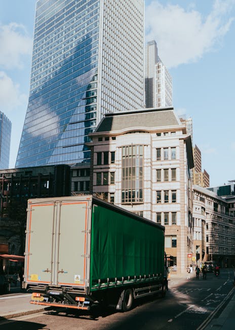 A large green moving truck with closed rear doors is parked on a city street in front of modern and historic buildings, including a tall glass skyscraper reflecting the blue sky and clouds. The truck is ready for a home relocation or furniture transport, typical of professional removals services such as those offered by Man with Van St Lukes. The urban environment features a paved road with pedestrian crossings, and some pedestrians are visible near the truck. The scene captures the loading process where moving vehicles are positioned for efficient logistics during a furniture or belongings transfer, with the background showcasing a mix of contemporary and older architecture, emphasizing a professional move within the city.