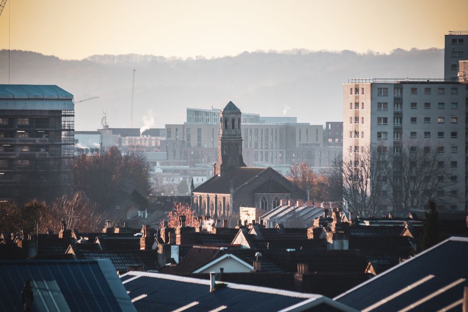 A cityscape view featuring a mix of residential and commercial buildings with rooftops in the foreground, constructed mainly of dark tiles and bricks. Behind the rooftops, there is an older church with a tall, pointed tower and arched windows, surrounded by leafless trees. In the background, modern high-rise buildings and a distant construction crane are visible, with some smoke or steam rising from structures in the mid-ground. The scene appears to be captured during daylight with soft natural lighting, highlighting the urban environment suitable for a home relocation or furniture transport context. This image is related to house removals and moving logistics, as from Man with Van St Lukes, supporting the topic of packing and loading processes involved in house removals in the Islington area.