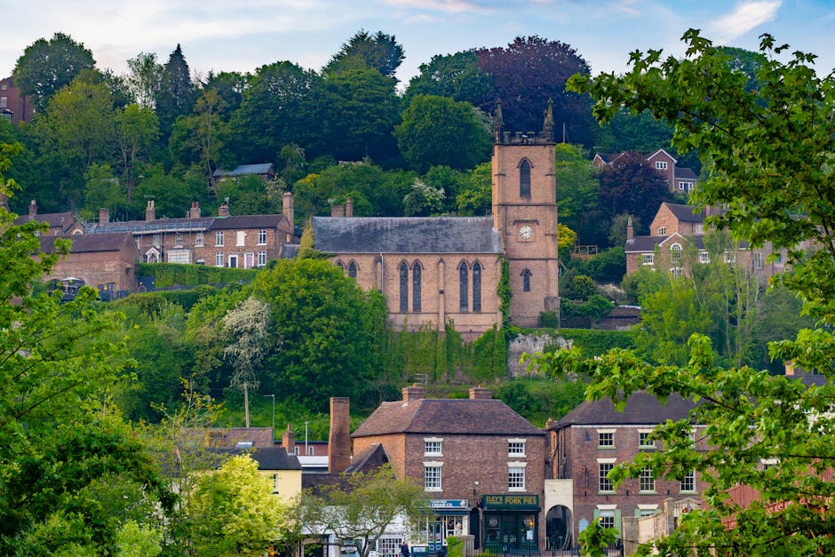 The image shows a picturesque residential area set on a hillside with a historic stone church featuring a tall clock tower situated among lush green trees and residential houses with brick facades and chimneys. The foreground includes several suburban homes, some with shopfronts, surrounded by vibrant foliage and trees, indicating a peaceful, well-established neighbourhood. The scene is illuminated with natural daylight, highlighting the various building materials such as brick and slate, and the greenery from trees and bushes that frame the view. This setting illustrates the typical environment encountered during house removals and home relocations, where careful furniture transport and packing are essential to navigate the hilly terrain and narrow pathways. Man with Van St Lukes specializes in efficient and damage-free removals, often involving complex loading processes and the use of equipment like trolleys and blankets to protect furniture during transport, especially in areas with historic architecture and landscaped surroundings.