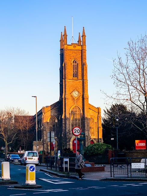 A historic stone church with a tall, rectangular clock tower featuring narrow, arched windows and a clock face near the top, illuminated by natural daylight with a clear blue sky background. In the foreground, a street scene includes parked cars, a person walking on the pavement, and traffic signage, indicating an urban setting. The church appears to be situated on a corner, with trees and street lamps nearby. This scene relates to home relocation or moving services provided by Man with Van St Lukes, which involves logistics around transporting household items to or from the church area, possibly during a house removal or local moving process.