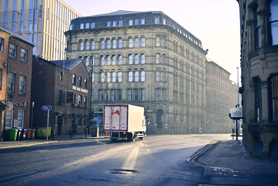 A white moving van from Man with Van St Lukes is parked on a narrow urban street lined with multi-storey residential buildings made of brick and concrete. The van is positioned close to a building with a pinkish facade, and its back doors are closed, ready for loading or unloading. The surrounding area includes parked cars, a discarded black trash bin, and a few other vehicles further down the street. The street surface is asphalt with visible yellow road markings, and the scene is illuminated by natural daylight, with the sky partly cloudy. The image captures the typical environment of a house or apartment relocation in St Luke's, showing the logistics involved in furniture transport and packing during a same-day move.