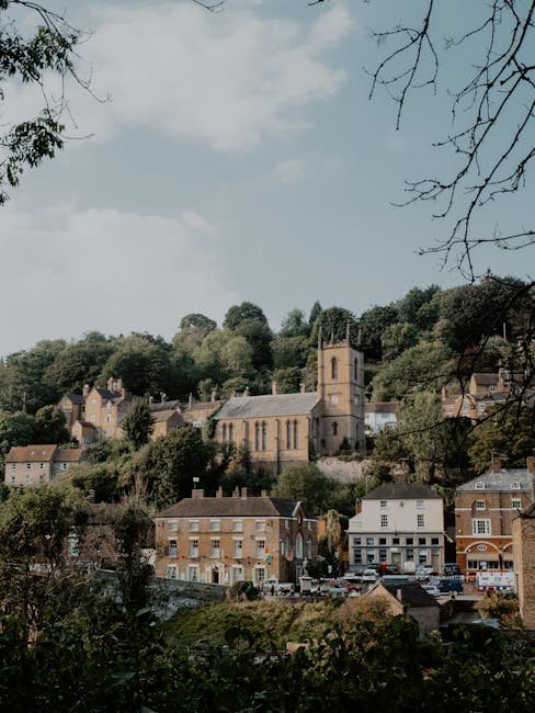 A scenic view of a small town with a prominent church featuring a square bell tower situated on a hillside. Surrounding the church are residential buildings and commercial shops at the base of the hill, with a densely wooded area behind the church. The sky above is partly cloudy with patches of blue, and the foreground includes tree branches and foliage. The area appears calm, with no people or vehicles visible, reflecting a typical residential neighbourhood that may require local house removals or furniture transport services for relocation purposes, as referenced in the context of St Luke's Guide: Parking & Access on Bunhill Fields Rd. This setting showcases the picturesque environment where home relocation or packing and moving activities could take place, highlighting the importance of careful planning for parking and access when conducting a move in such scenic locations.