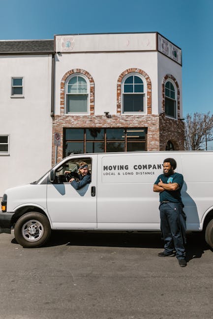 A white moving van with the label 'MOVING COMPANY LOCAL & LONG DISTANCE' parked in front of a two-storey residential building. Two men are engaged in a conversation beside the van; one is seated inside the driver’s seat, wearing sunglasses and a checked shirt, while the other, standing outside, has arms crossed and is dressed in a dark shirt and pants. The building features a brick facade on the lower level with two large windows, and a white upper level with two arched windows and additional artwork or signage on the top corner. The scene is set on a clear day with a blue sky, and the men appear to be discussing or coordinating efforts related to home relocation or furniture transport, typical of professional removals services by [COMPANY_NAME].