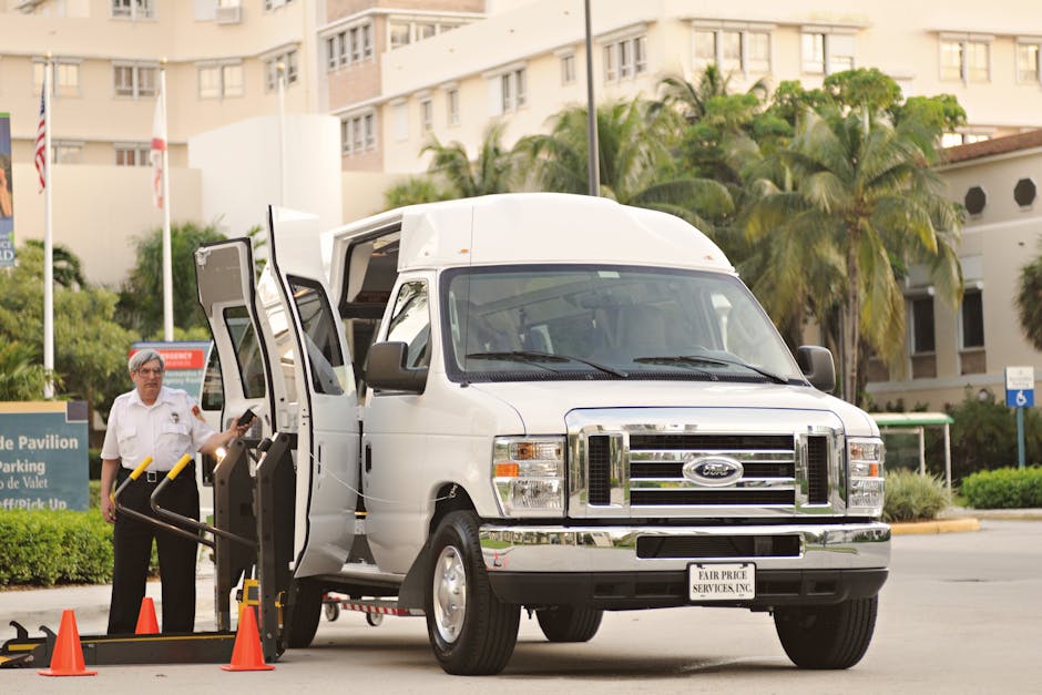 A white moving van with the label 'MOVING COMPANY LOCAL & LONG DISTANCE' parked in front of a two-storey residential building. Two men are engaged in a conversation beside the van; one is seated inside the driver’s seat, wearing sunglasses and a checked shirt, while the other, standing outside, has arms crossed and is dressed in a dark shirt and pants. The building features a brick facade on the lower level with two large windows, and a white upper level with two arched windows and additional artwork or signage on the top corner. The scene is set on a clear day with a blue sky, and the men appear to be discussing or coordinating efforts related to home relocation or furniture transport, typical of professional removals services by [COMPANY_NAME].